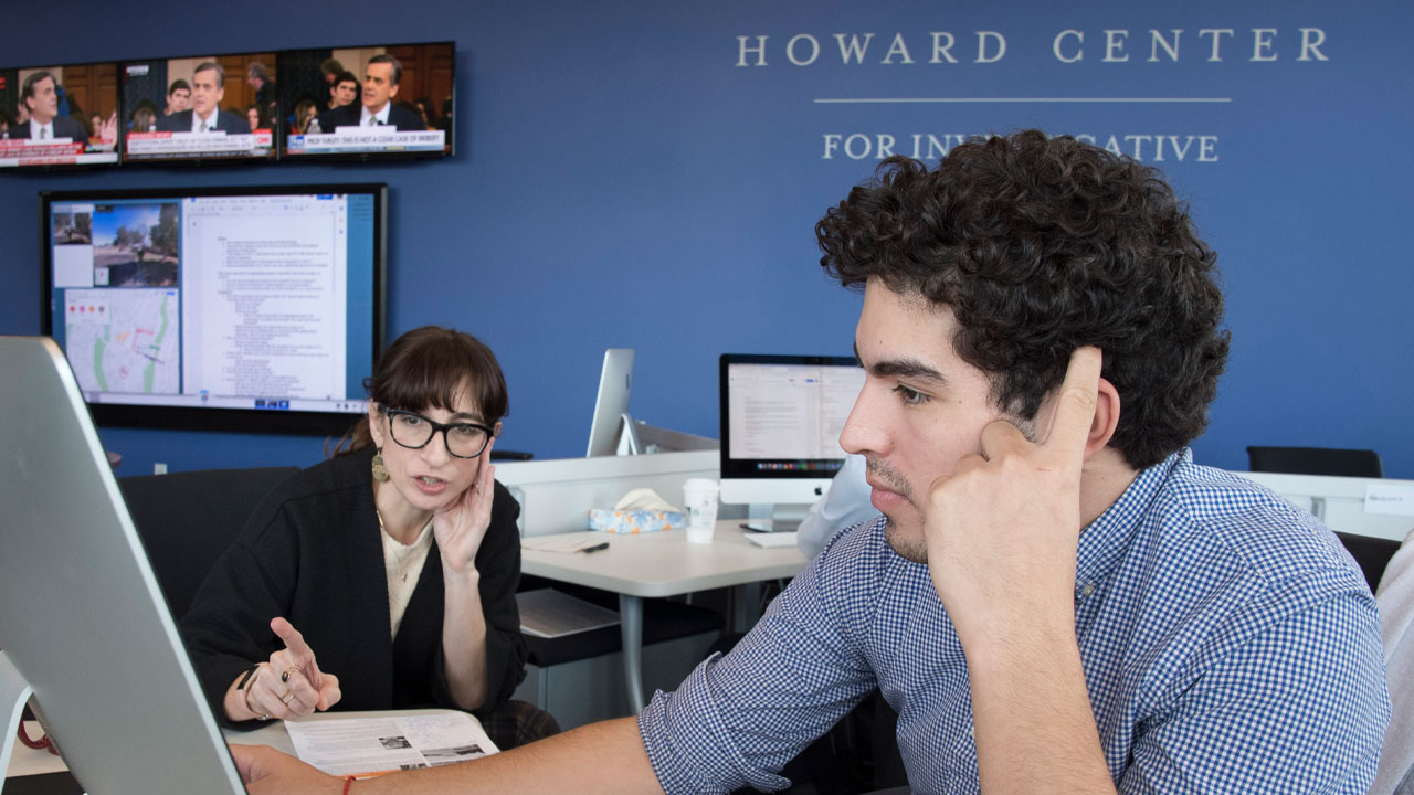 Two people discussing at desks in the Howard Center with multiple computer monitors and news screens.