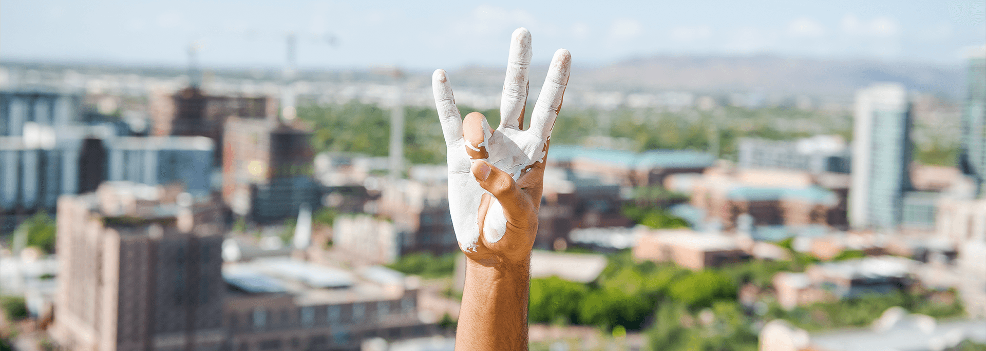A hand covered in paint holding up the Sun Devil pitchfork sign in front of a city background.