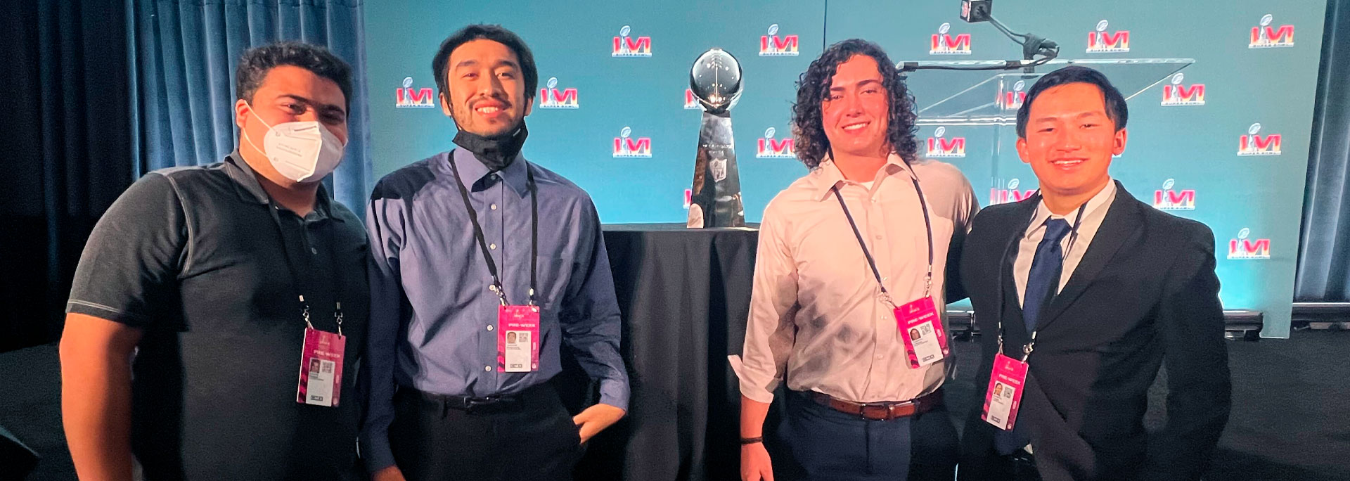 Four individuals in business attire standing in front of a backdrop with microphones and a Super Bowl trophy.