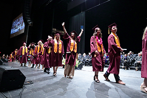 Graduates in maroon caps and gowns walk across a stage during a ceremony.