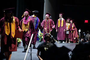 Graduates in maroon caps and gowns walk across a stage during a ceremony.