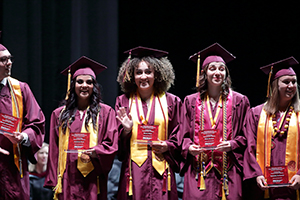 Five graduates in maroon caps and gowns holding red plaques on a stage.