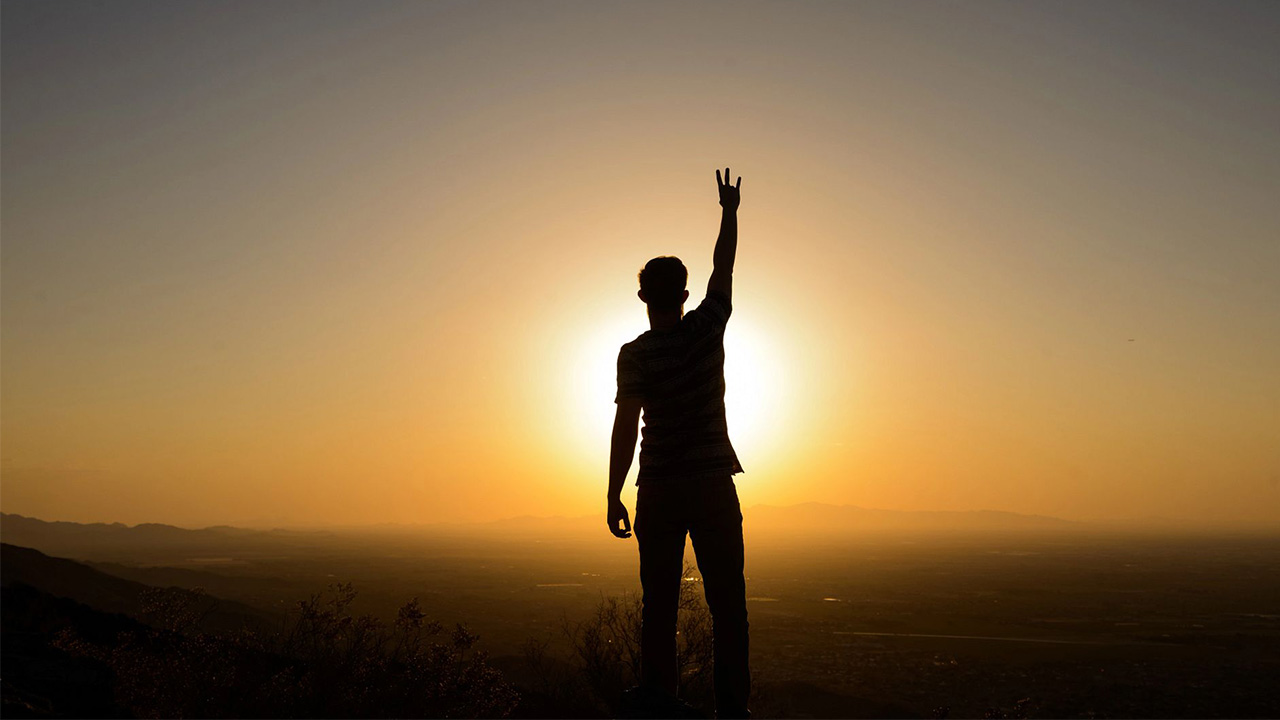 An ASU student holds up the pitchfork hand sign in front of a sunset.
