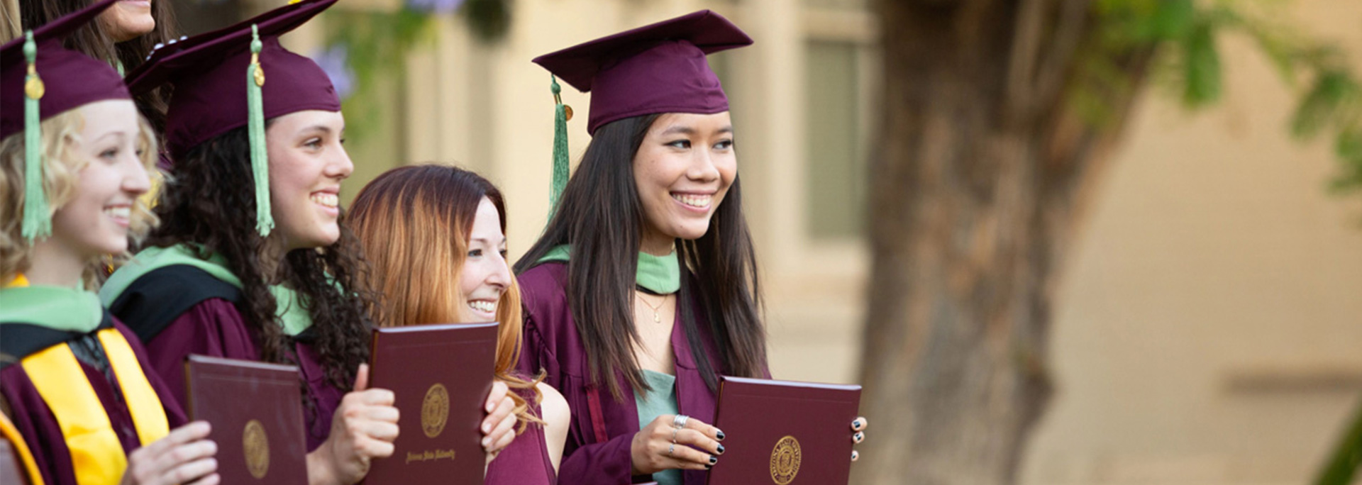 
		A group of new graduates wear maroon caps and gowns while holding their ASU diplomas.		