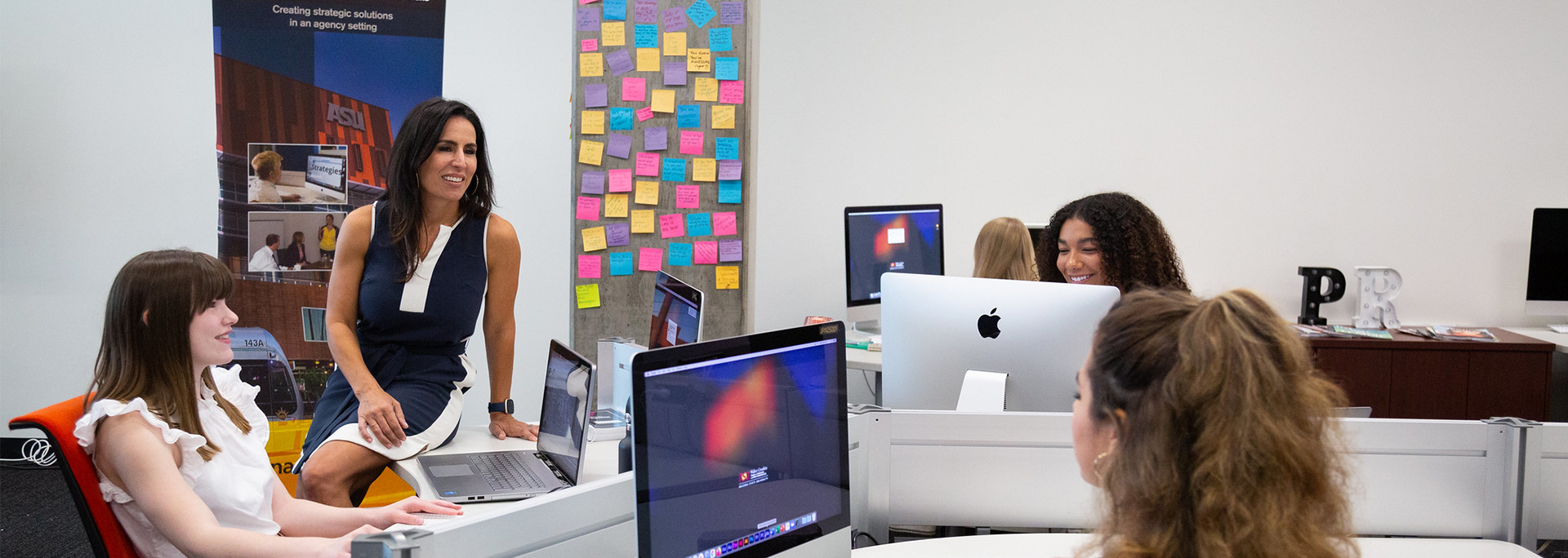 Three students in the Public Relations Lab talk with their professor seated at their desks.