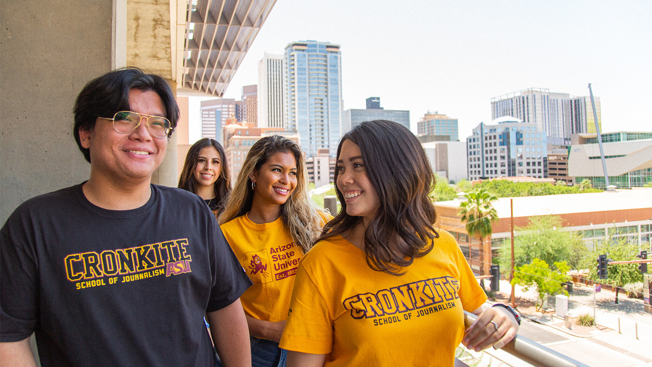 Four Cronkite students in ASU shirts stand on a balcony with Downtown Phoenix in the background.
