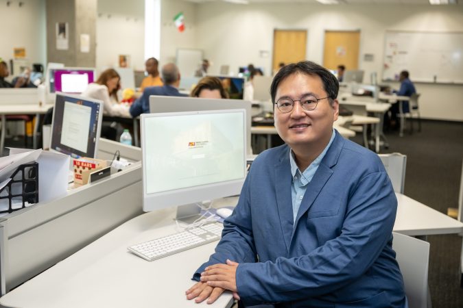 Tag Lee wears a blue blazer and sits in front of a newsroom computer